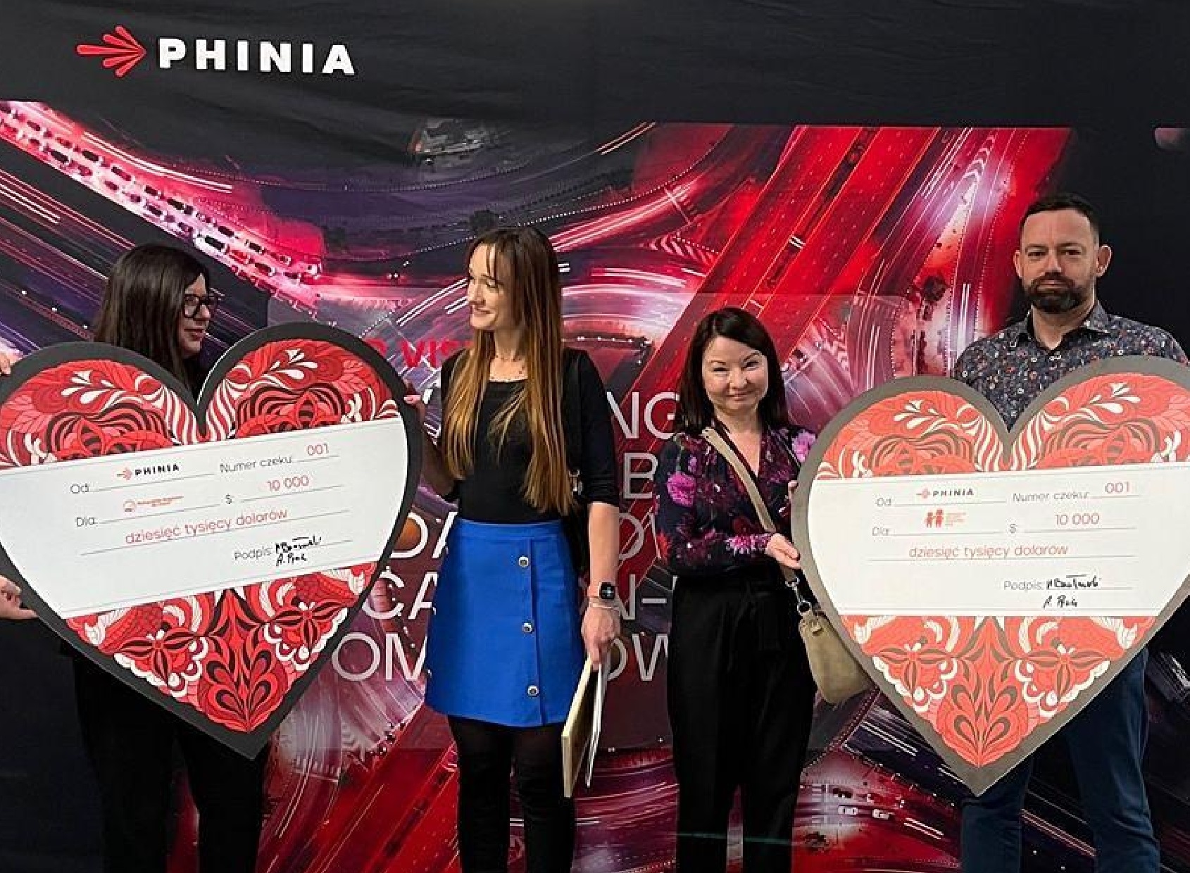 Group of employees standing in front of a PHINIA charity event backdrop, holding large red heart-shaped donation cheques