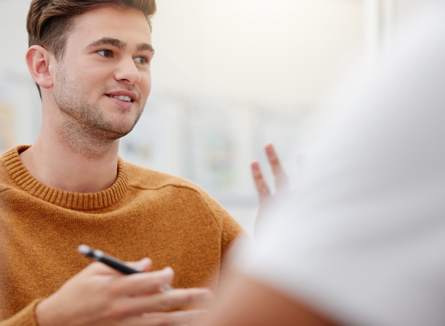 A young male engineer in a casual sweater talking to a colleague wearing a safety helmet inside a modern industrial workspace