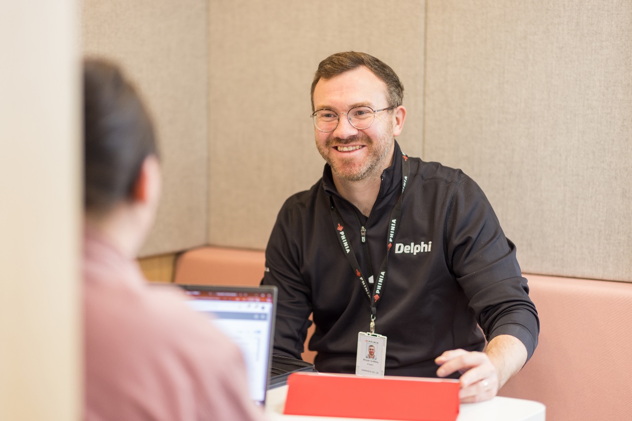 Man smiling in a business casual setting, sitting at a desk during a meeting with a laptop and tablet nearby