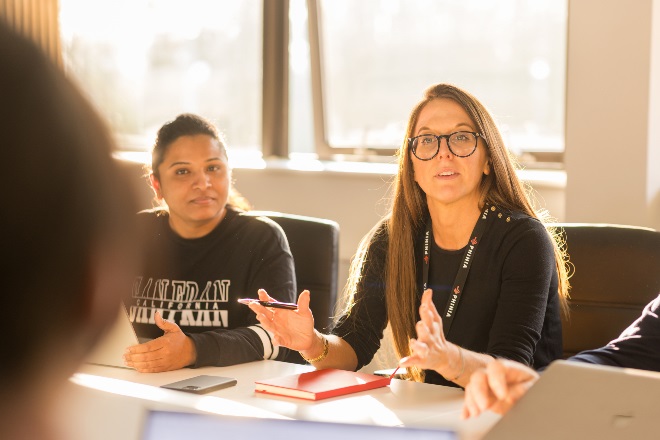 Woman leading a discussion in a bright meeting room, gesturing while speaking, with another woman listening attentively beside her.